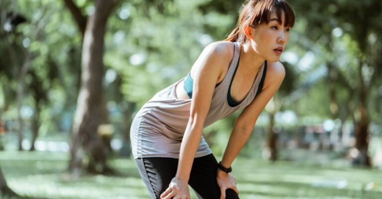 Recovery - Tired ethnic female in sportswear looking away while leaning on knees having rest after workout in park on sunny summer day